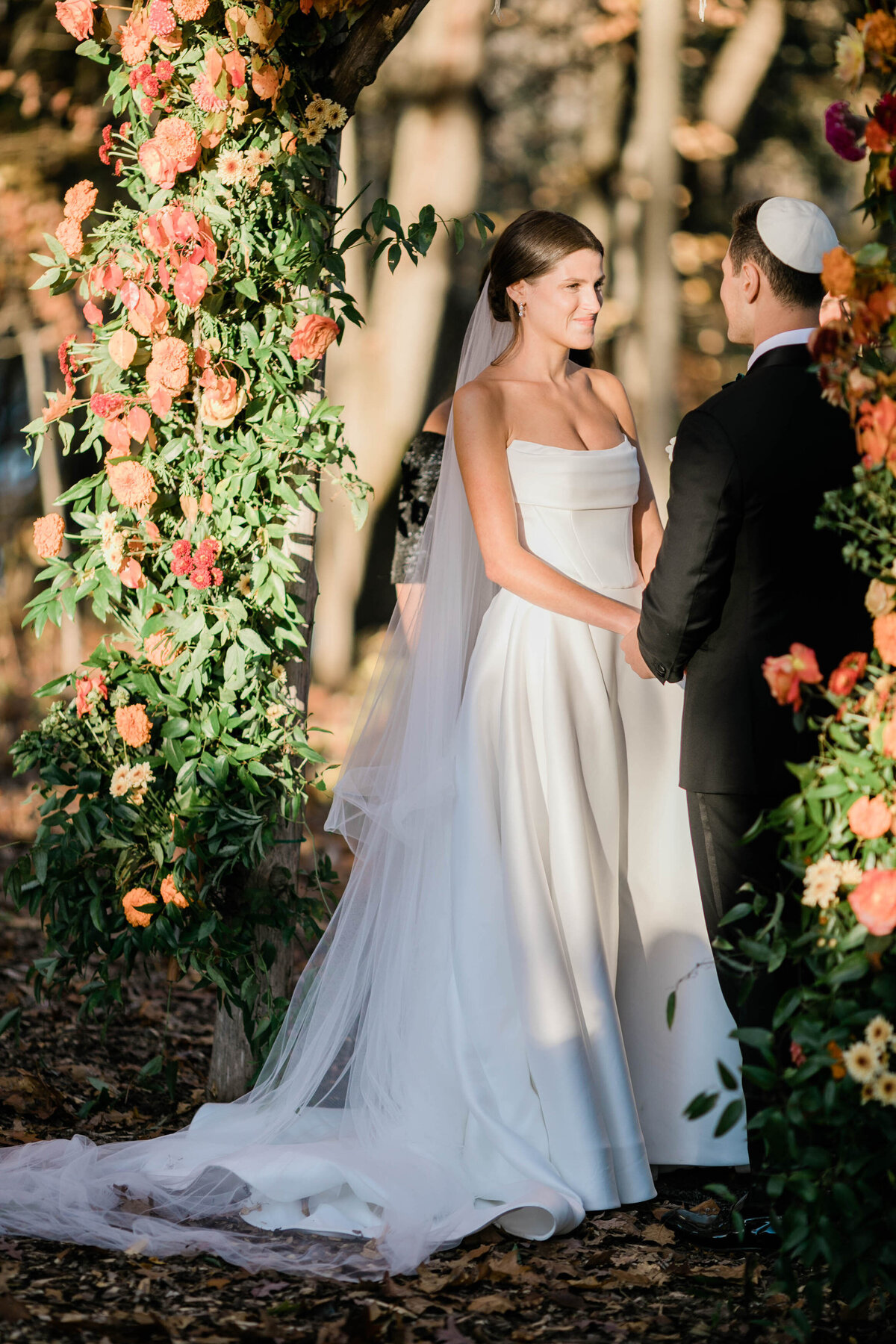 The bride, in a flowing white gown with a long veil, gazes lovingly at her groom as they exchange vows under the floral chuppah