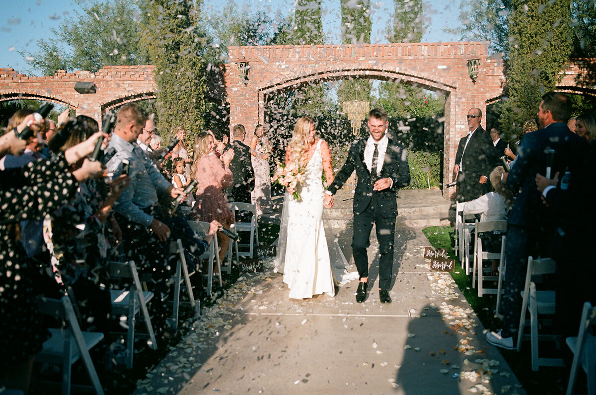 Newlywed couple exiting their wedding ceremony while holding hands and walking through biodegradeable confetti