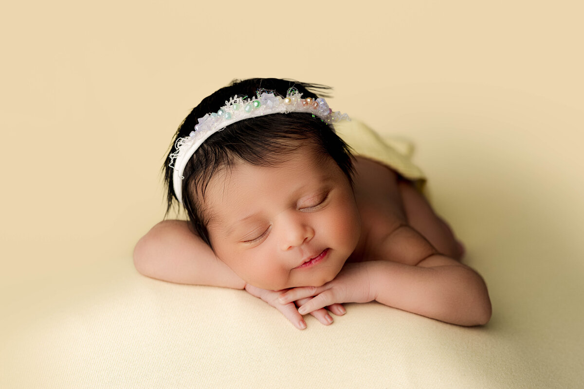 Newborn girl resting on a creamy yellow backdrop with hands folded under her chin and a delicate floral headband — warm and airy studio portrait