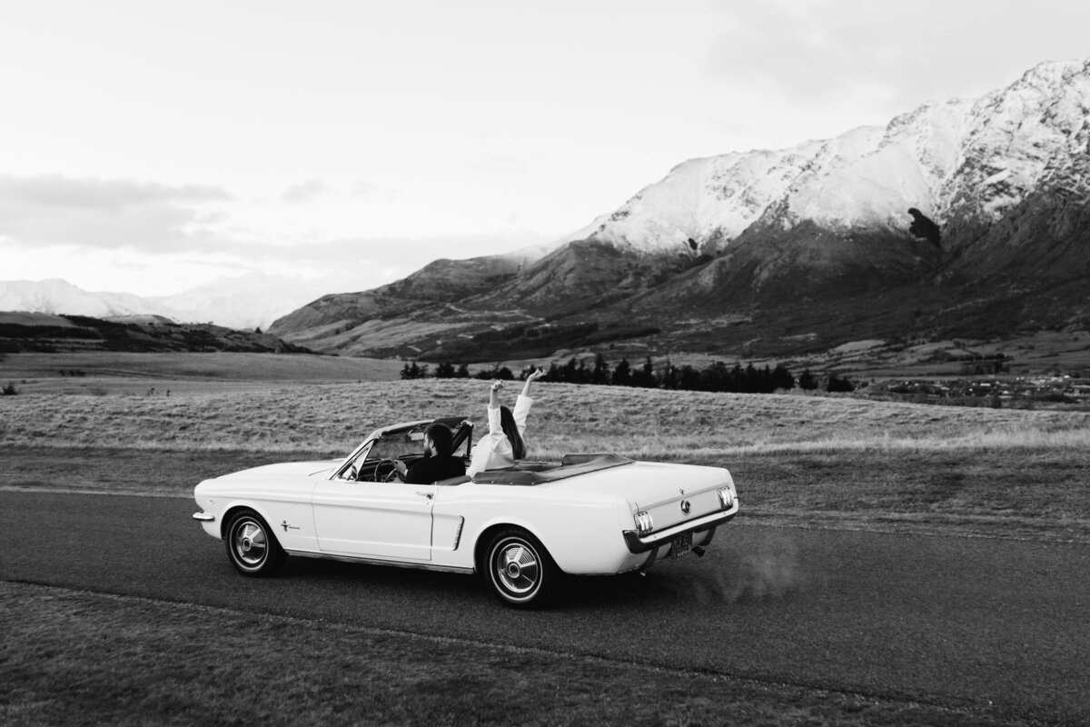 The Lovers Elopement Co - bride and groom drive through Queenstown mountain landscape in open top classic car at wedding elopement
