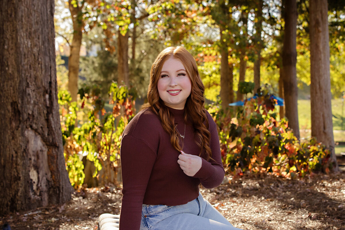 Portrait of a high school senior in cranberry shirt and jeans sitting on a bench in a South Charlotte park with beautiful golden fall foliage tones on an Autumn day