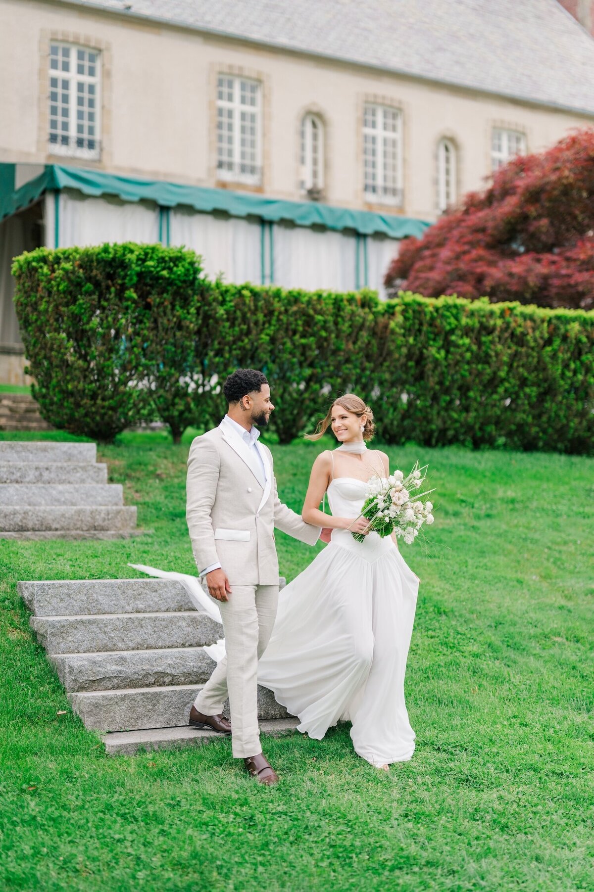 A bride holding hands with her groom and smiling at each other in front of Glen Manor House. 