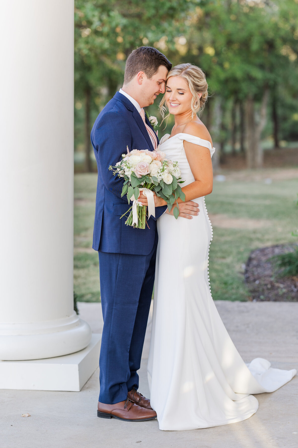 A bride and groom share a quiet embrace beside a stately white column at a wedding venue near Flower Mound, Texas. Her fitted off-the-shoulder gown and soft bouquet of blush roses contrast beautifully with his navy suit in this elegant, southern-inspired moment.