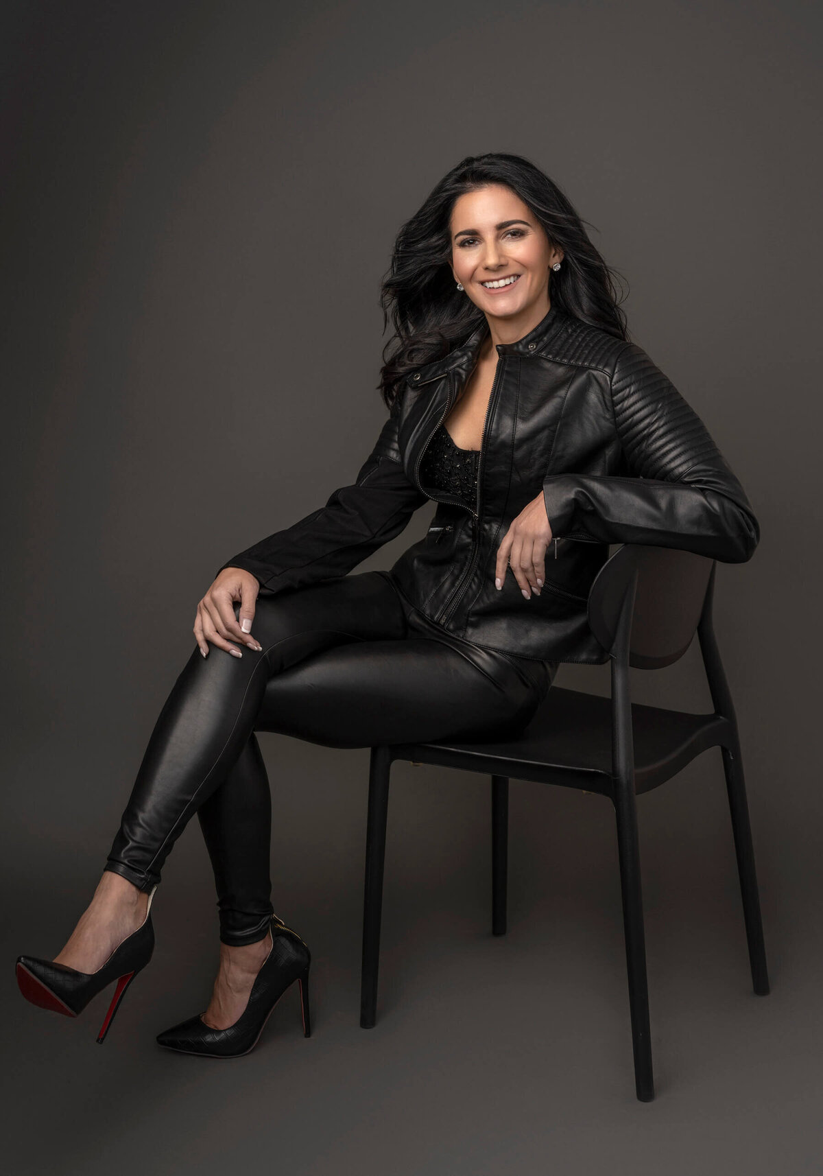 Black and white portrait of a woman in a leather jacket and leggings, sitting in a chair with a bold, confident smile.