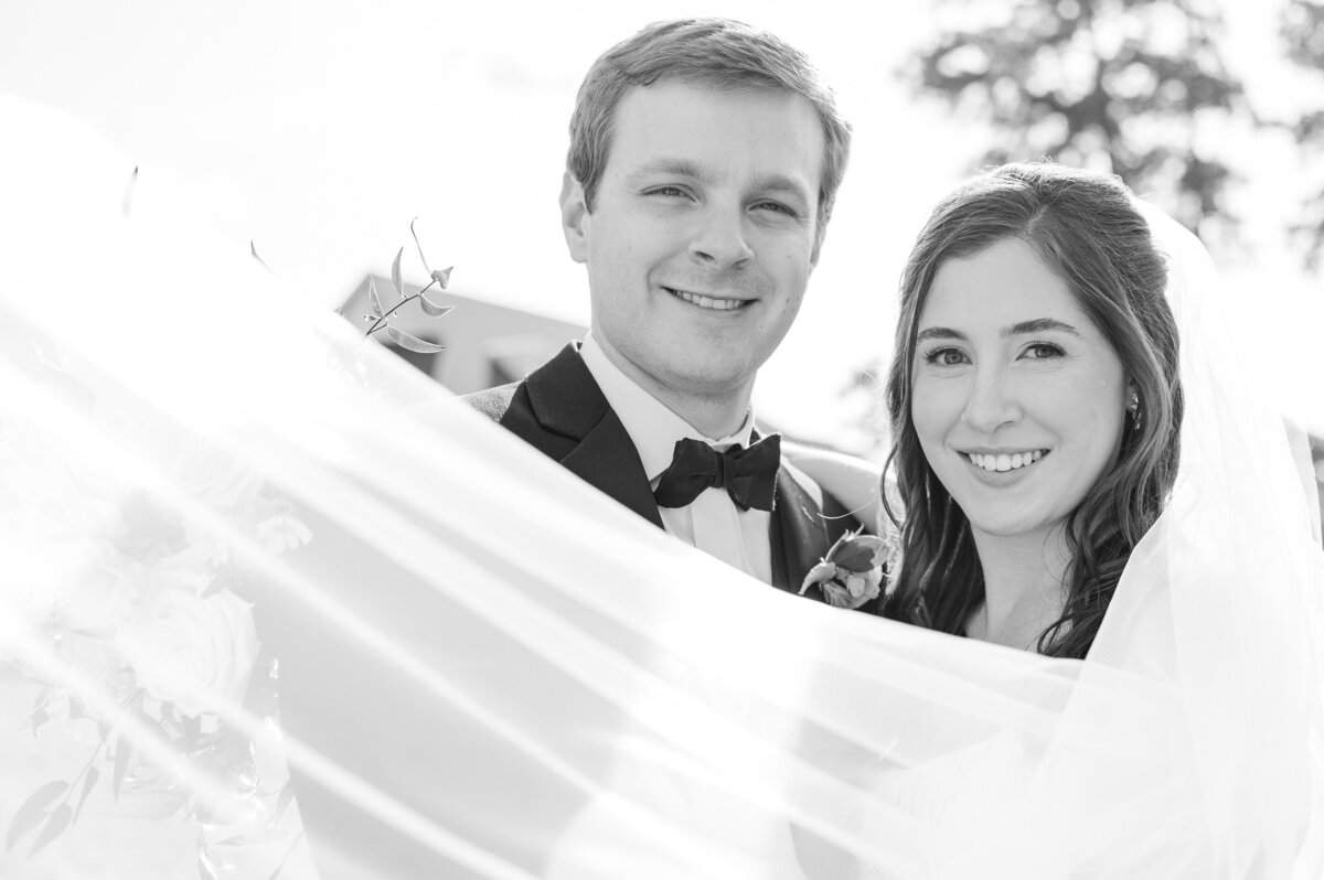 A white veil sweeping across the faces of a loving bride and groom at their wedding at The Prestonwood Country Club in black and white by JoLynn Photography, a Raleigh wedding photographer