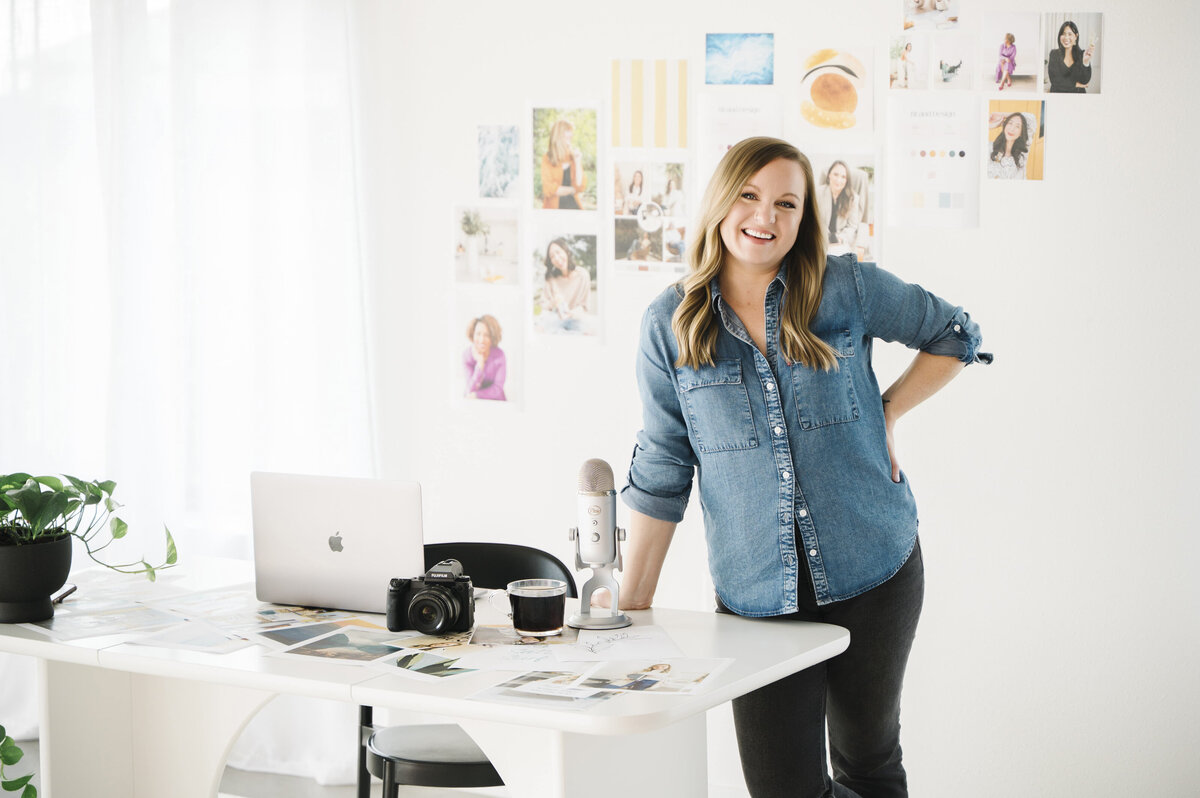 Brand photographer, Krista Marie, smiling at the camera and standing at her desk with a microphone, camera and laptop