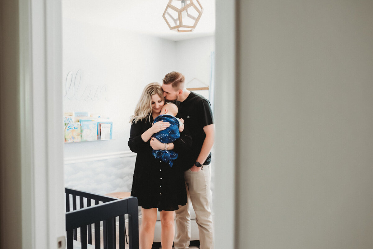 Mama holds baby boy while dad kisses mama, standing in nursery