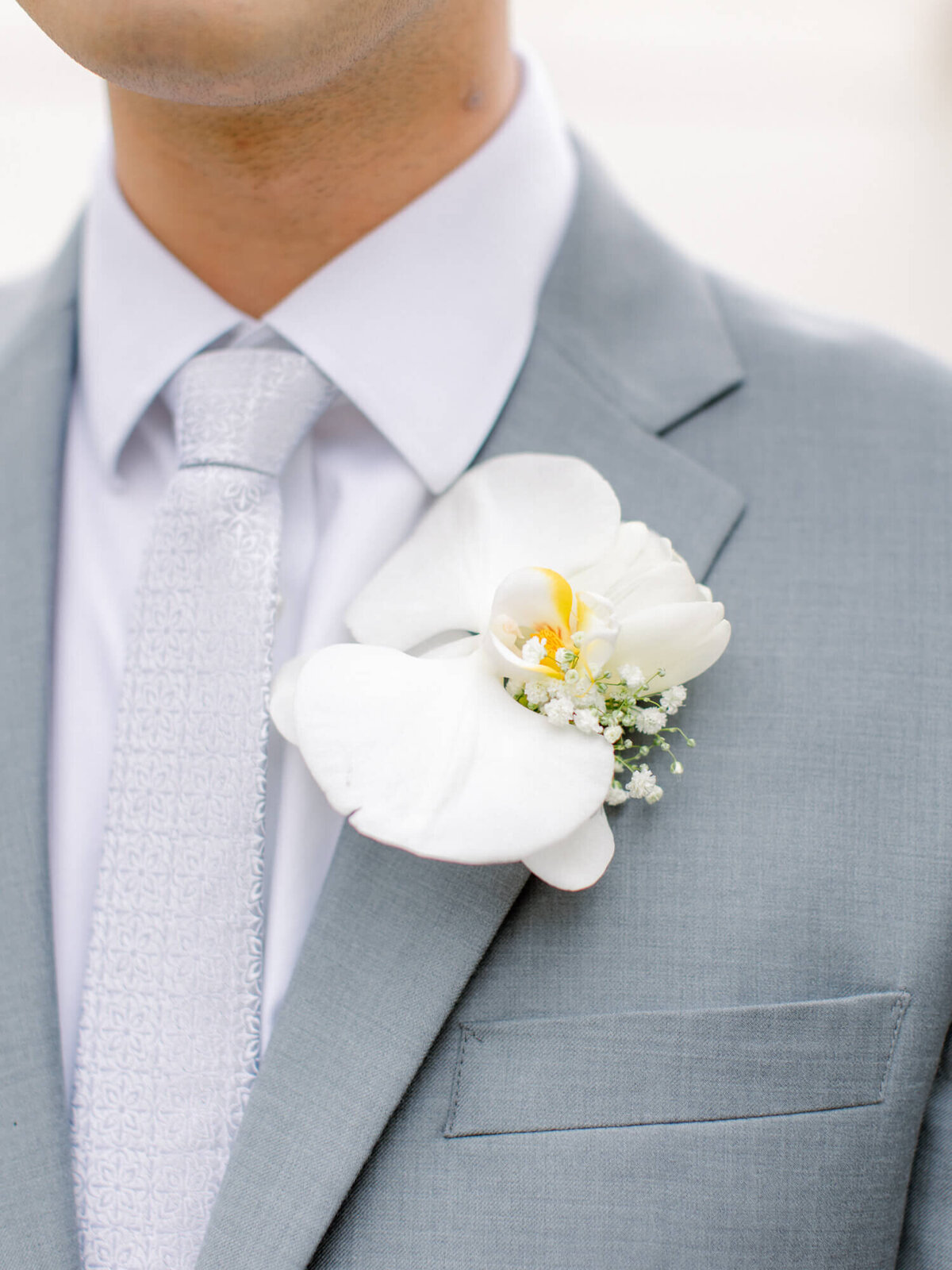 Close-up of a groom in a light gray suit with a white textured tie. A white orchid and baby's breath boutonniere is pinned to the suit, creating an elegant look.