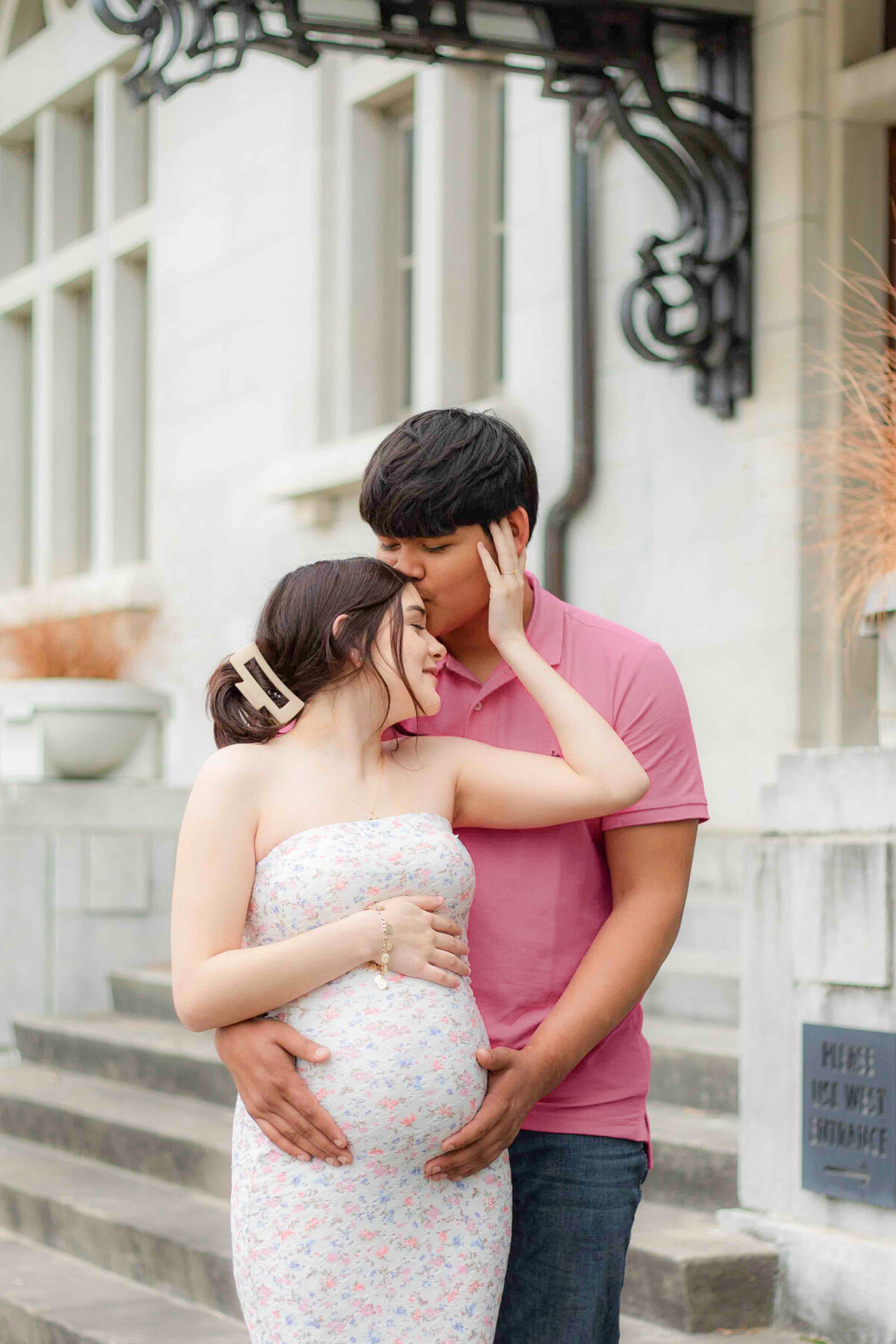 A romantic and intimate portrait of a pregnant mother standing in front of her partner, his lips softly pressed to her forehead as they cradle their growing baby bump together.