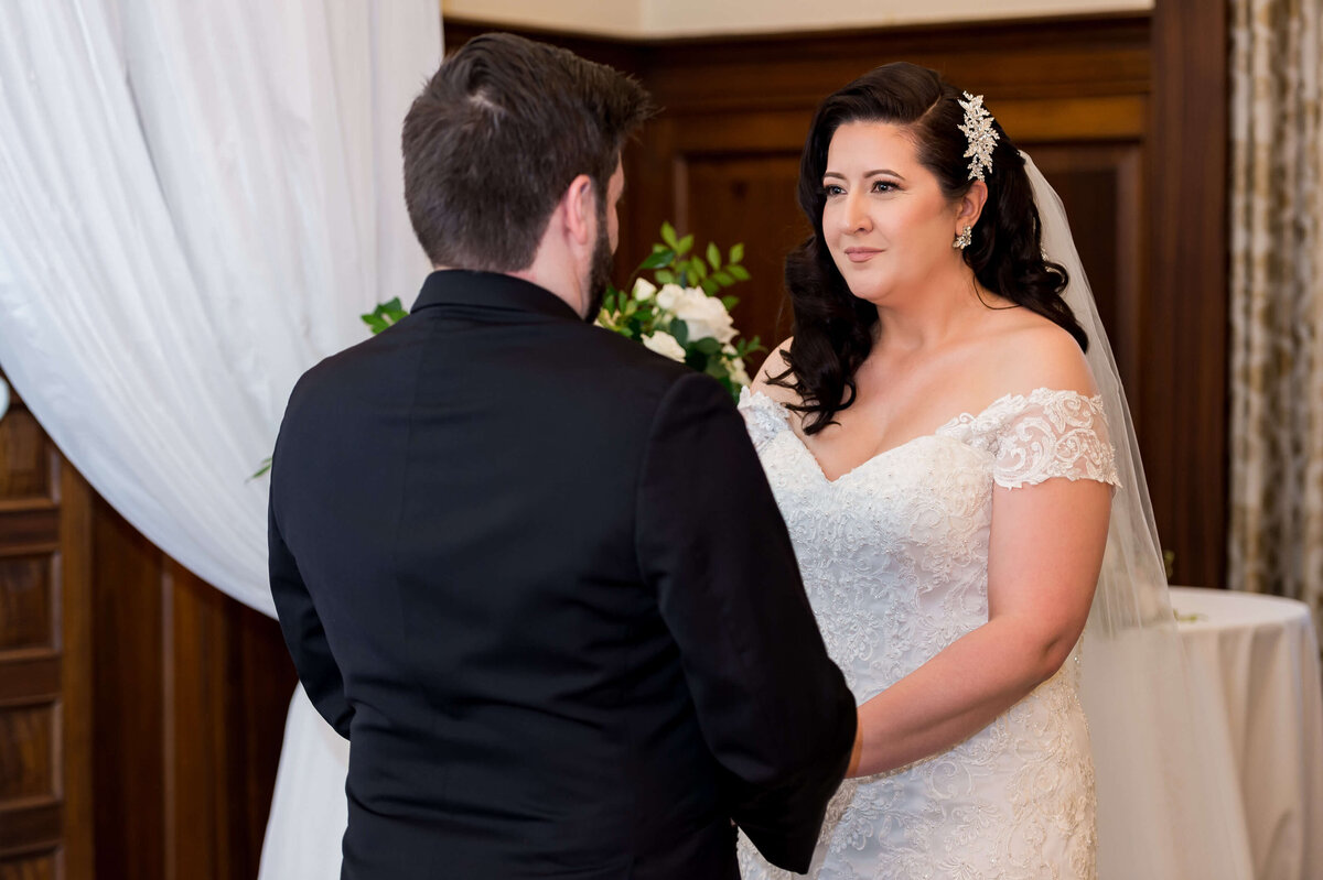 Ottawa wedding photography showing a bride smiling at her groom in a black tuxedo during their vows.  Taken inside at the Chateau Laurier hotel