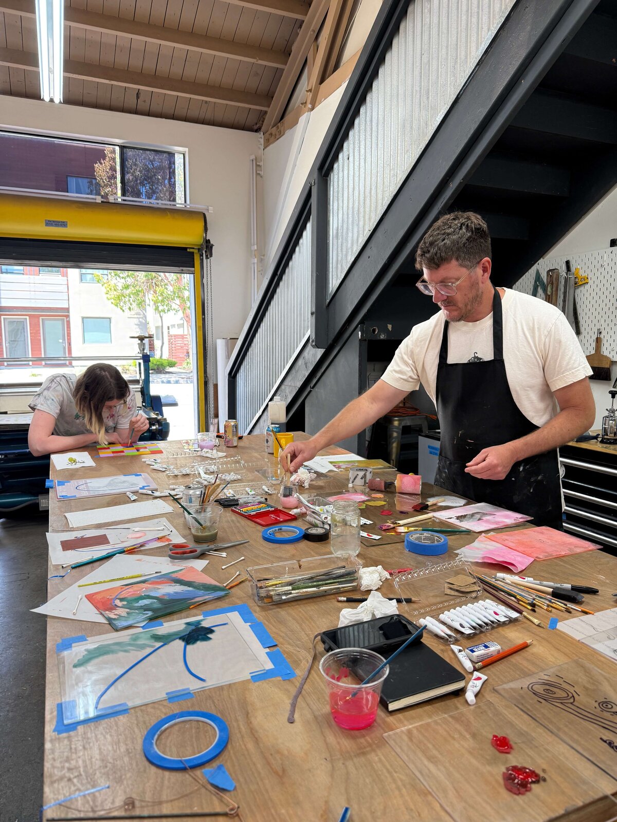 Two artists working side by side at a long shared table filled with monoprinting tools and printed works in progress.