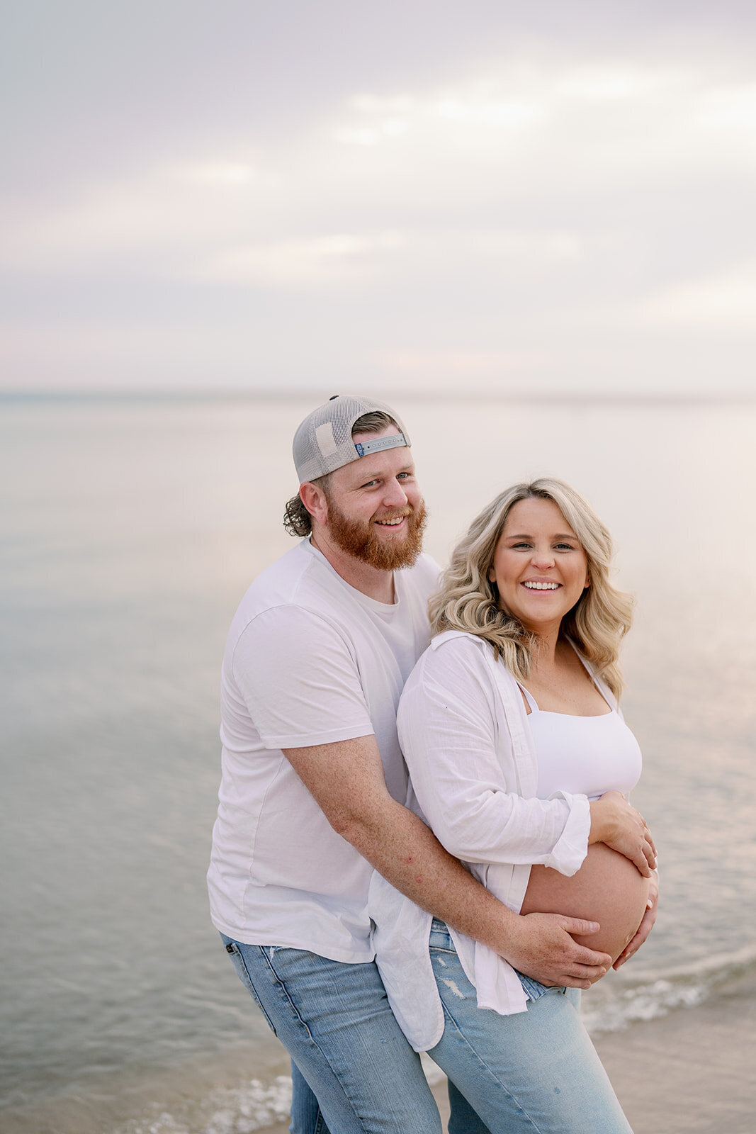 Expecting couple posing together on the shoreline at South Haven North Beach during their maternity photoshoot at sunset.