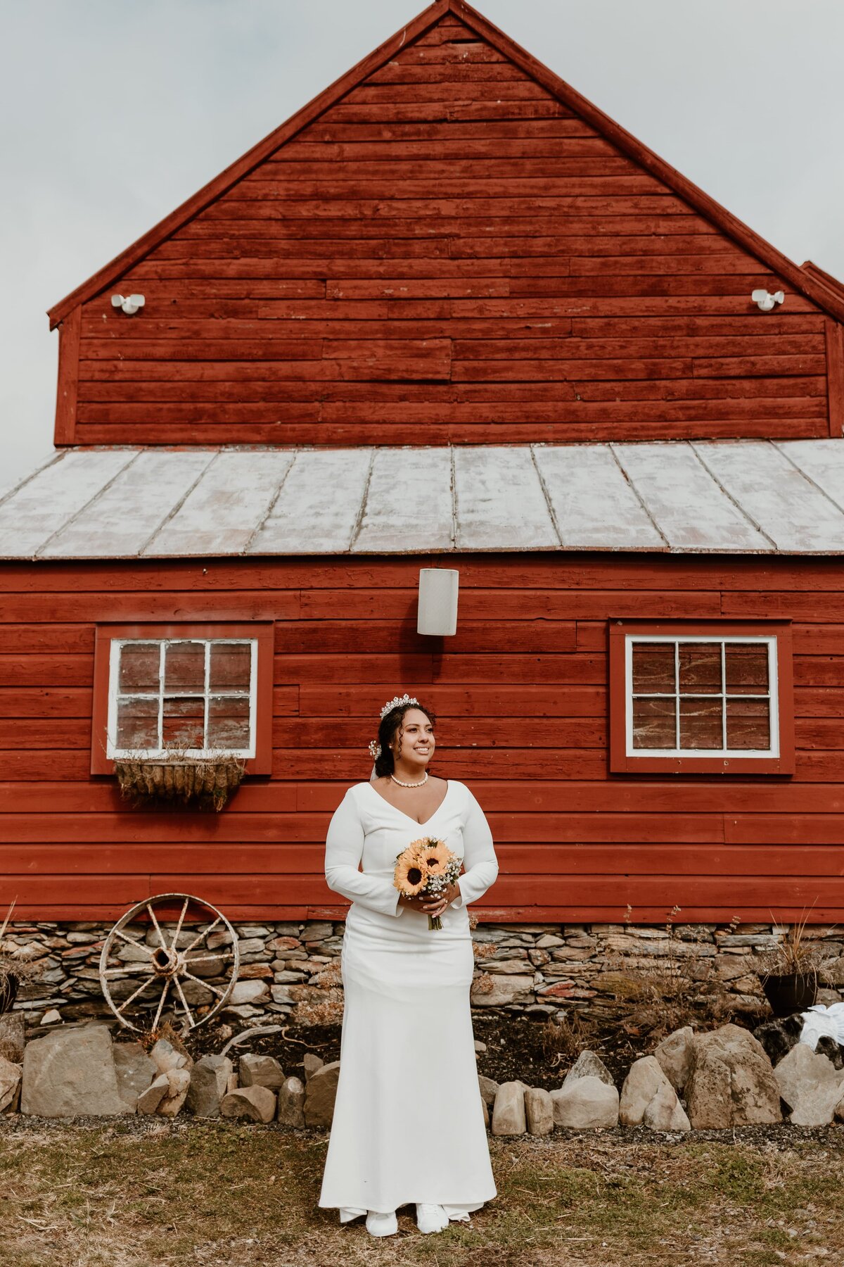 A smiling bride holding a bouquet of sunflowers stands in front of a rustic red barn at Black Snake Brewing Company on Old Adriance Farm. The barn features a quaint window with a flower box and a vintage wheel decoration to the side.