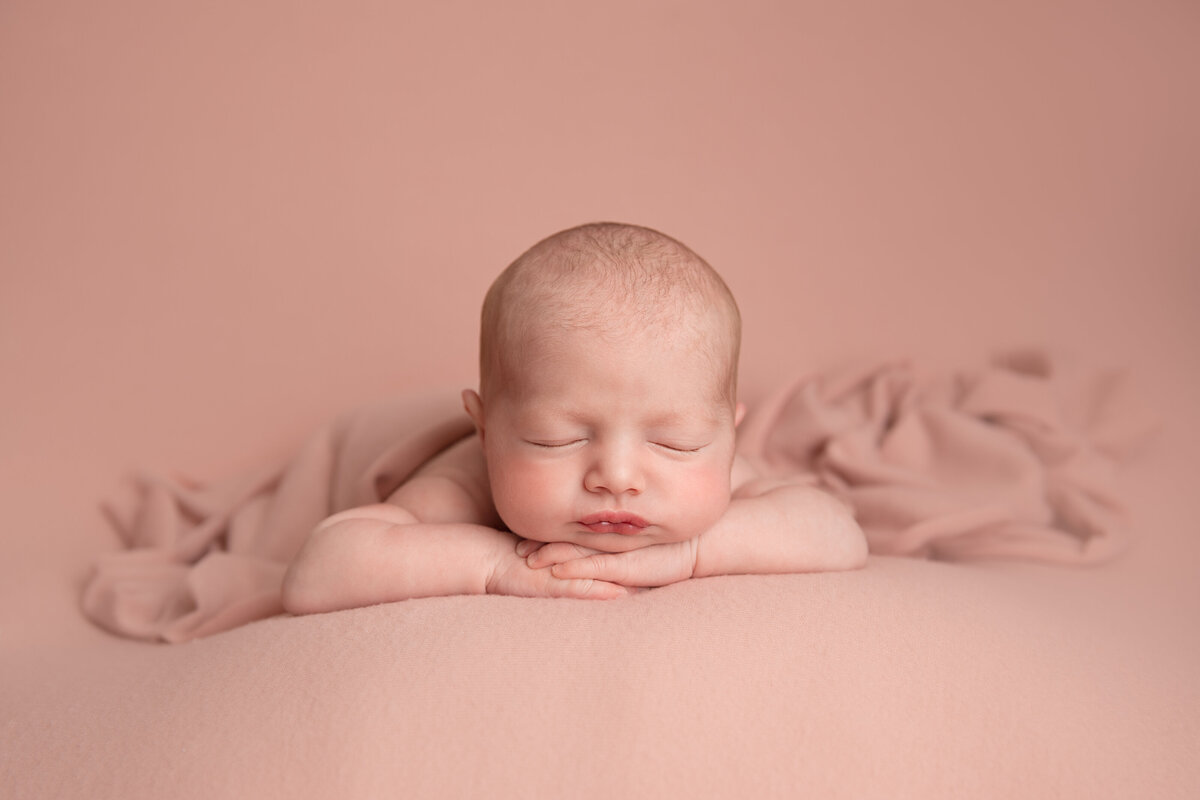 Newborn baby girl sleeping on her stomach and leaning forward on her hands on a light pink background.