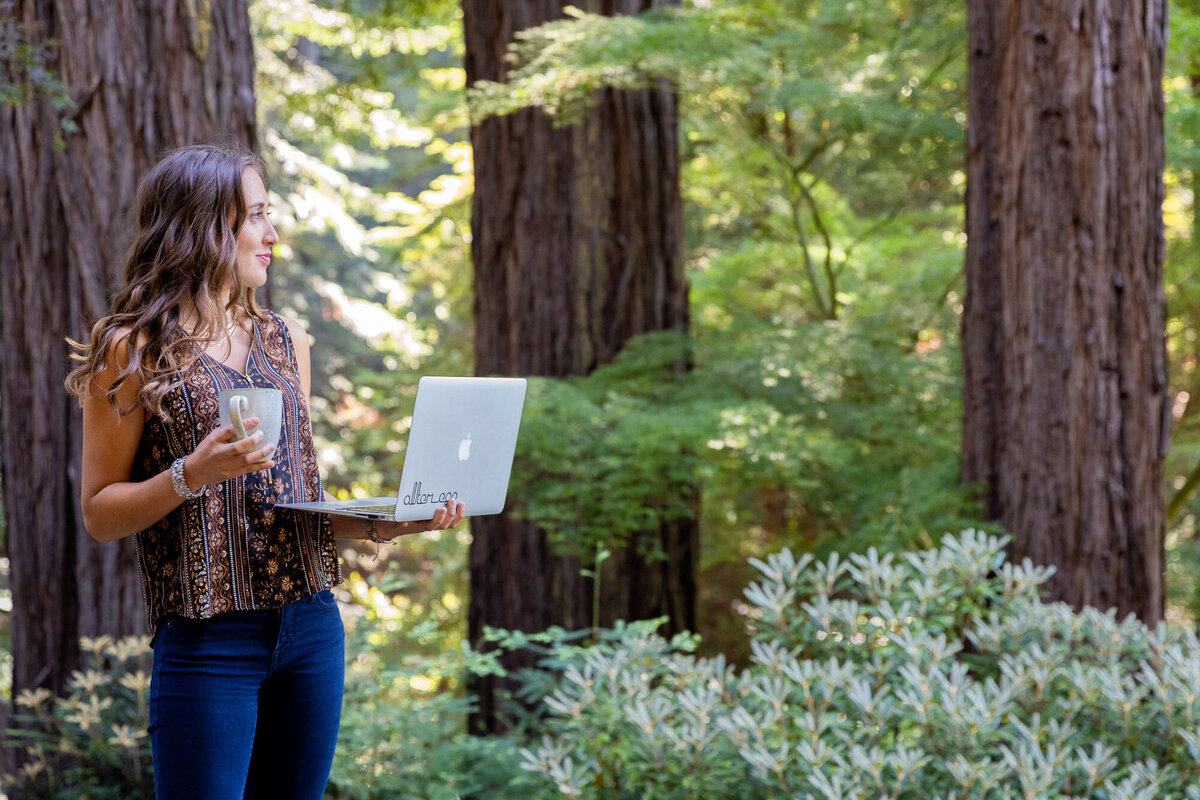 Cassandra outside holding laptop and coffee mug standing and looking out at the redwood tress