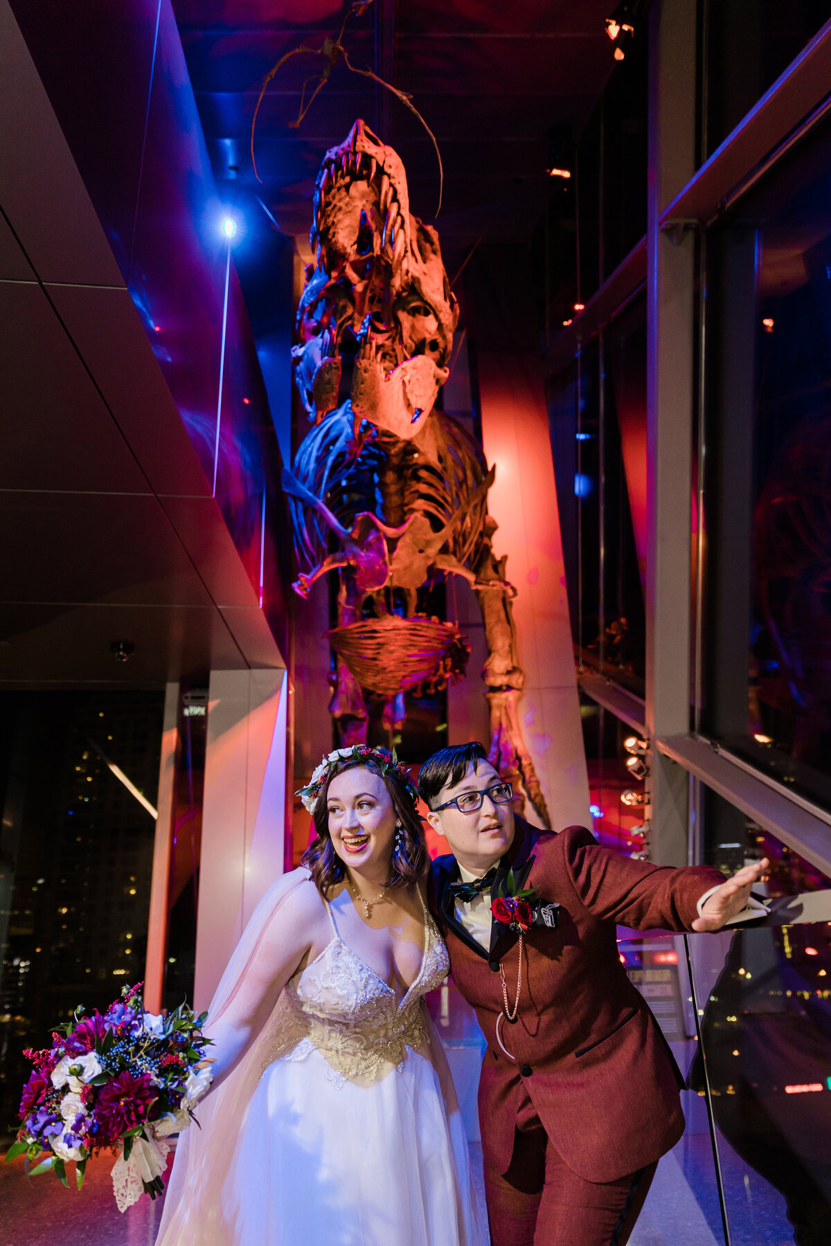 Portrait of two brides playfully posing underneath a tyrannosaur rex skeleton after their wedding at the Perot Museum of Nature and Science  in Dallas, Texas. The bride on the left is wearing a long, intricate, white dress with a flower crown and veil while holding a bouquet. The bride on the right is wearing a burgundy suit with a bowtie and boutonniere. The skeleton above them is bathed in colorful light.