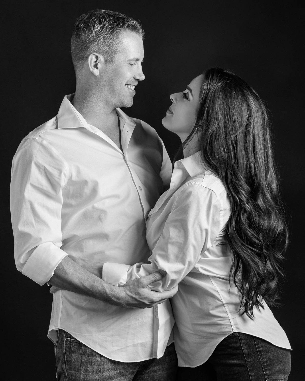 Black and white studio portrait of a couple holding each other and smiling warmly, photographed on a dark backdrop to highlight their connection.