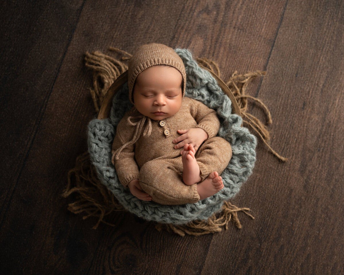 Newborn baby in tan knit bonnet and romper sleeping in a round bowl with layered textures on wood floor | Studio newborn session in Austin and Cedar Park
