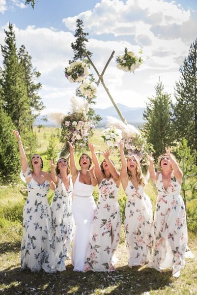 A bride and her bridesmaids dressed in floral dresses toss their bouquets into the air playfully.