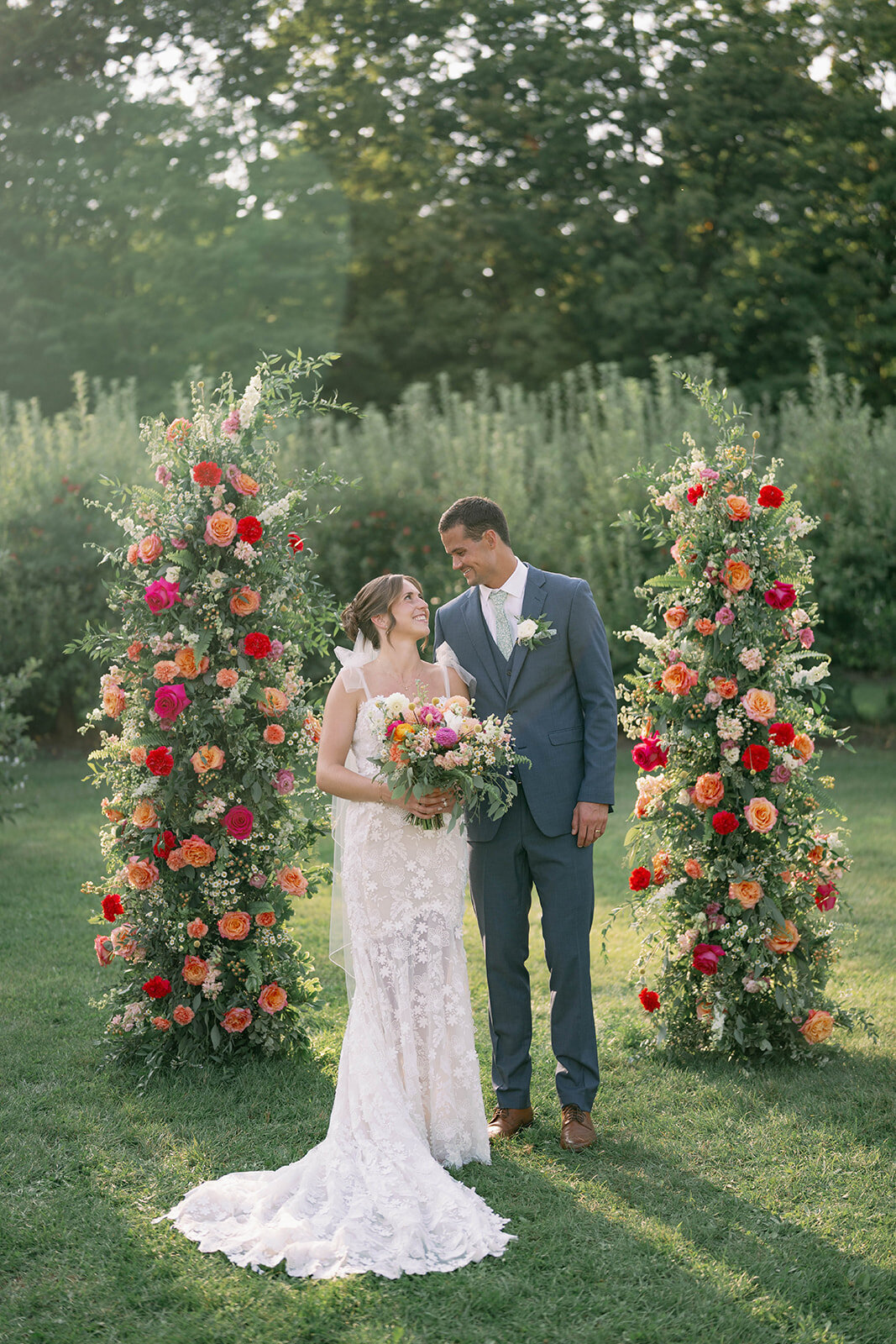 Bride and groom looking at each other while standing in front of their colorful floral arch at their orchard ceremony at The Cherry Barn in Frankfort MI.