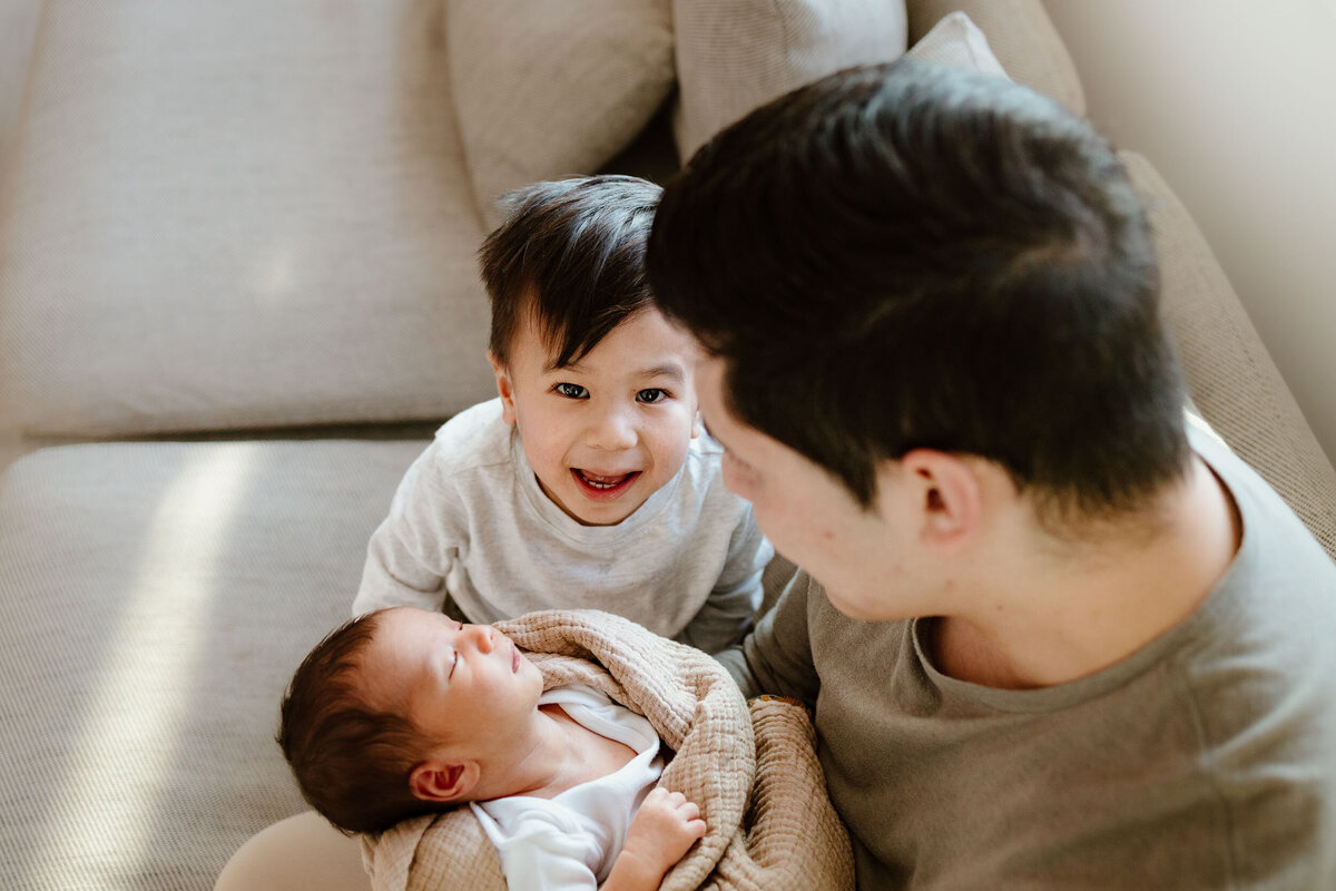 dad holding baby next to brother sitting on couch