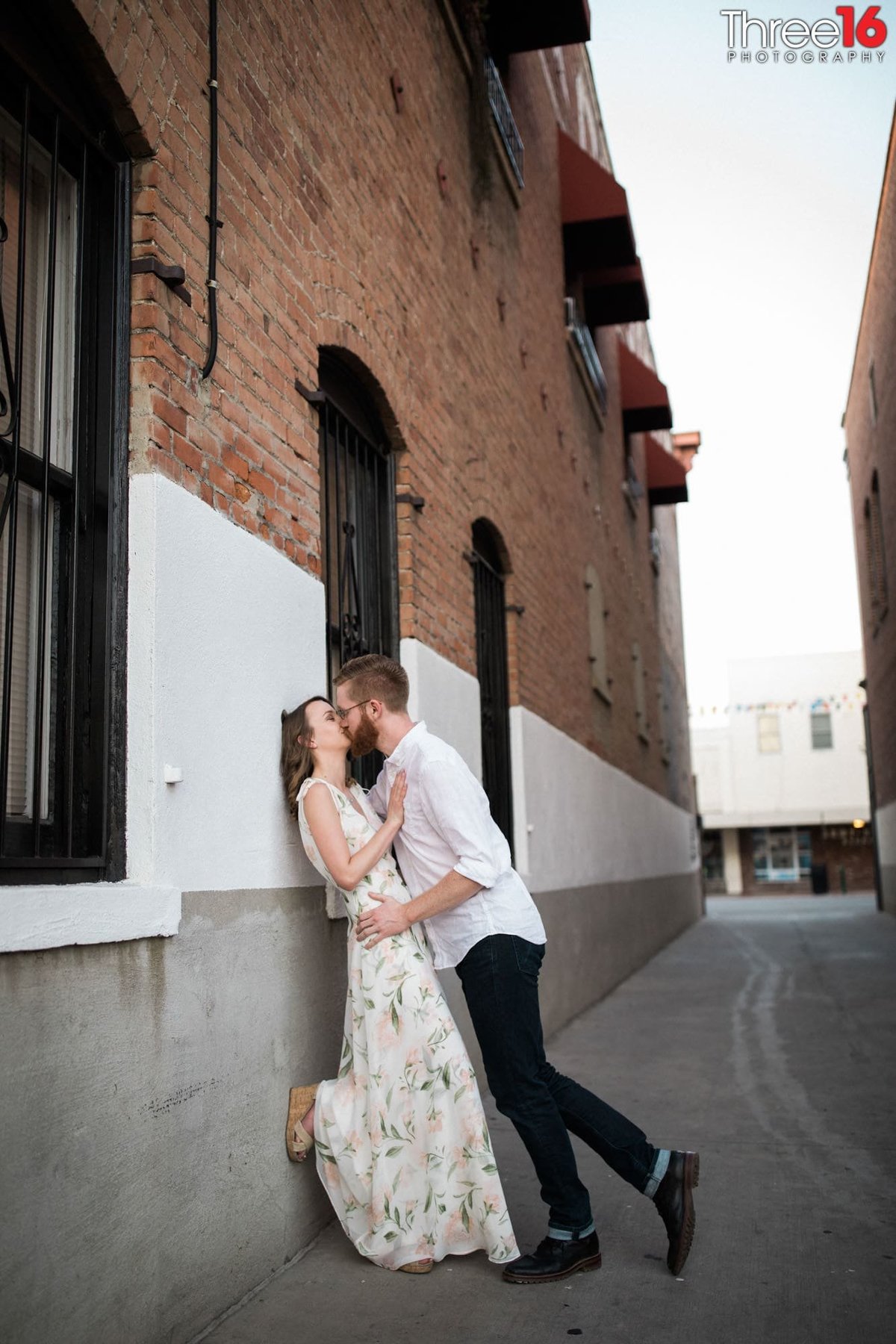 Groom to be leans into a kiss as his Bride is against a brick wall