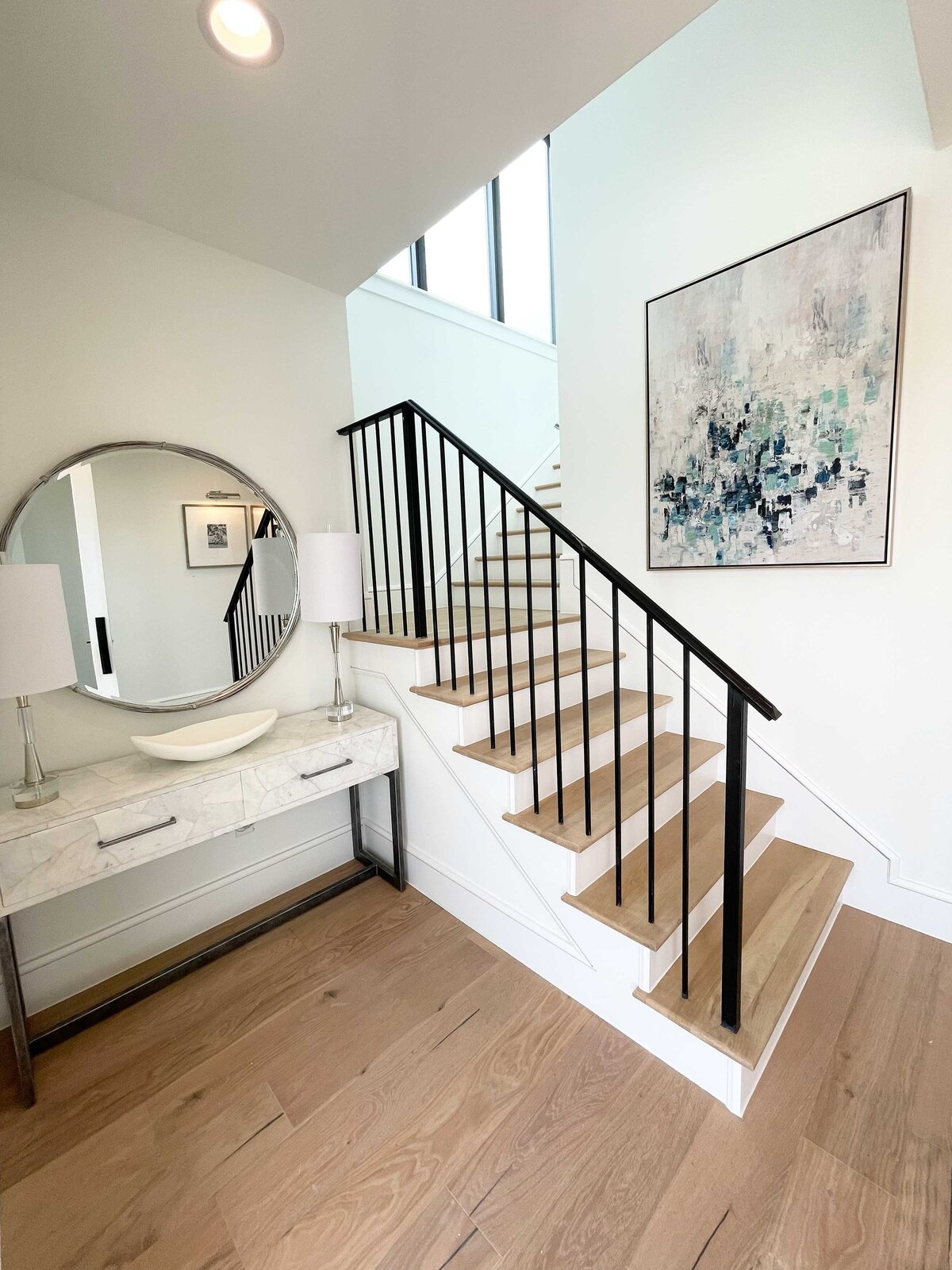 Expert home interior design featuring a peaceful house hall with pastel colors, white lamps, a circular mirror, and artwork above the stairs to create a serene and elegant atmosphere.