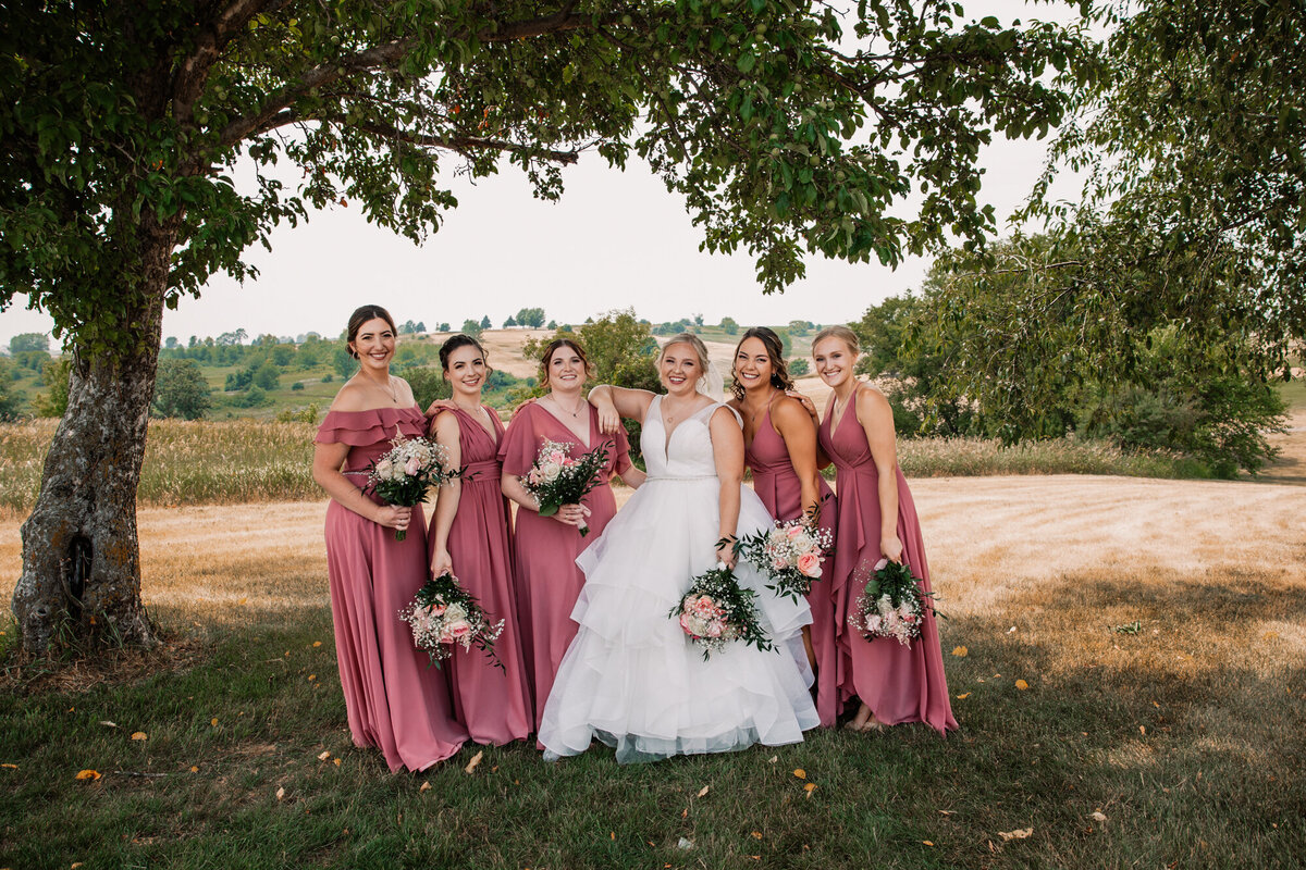 bride stands with smiling bridemaids in pink under a tree in the grass