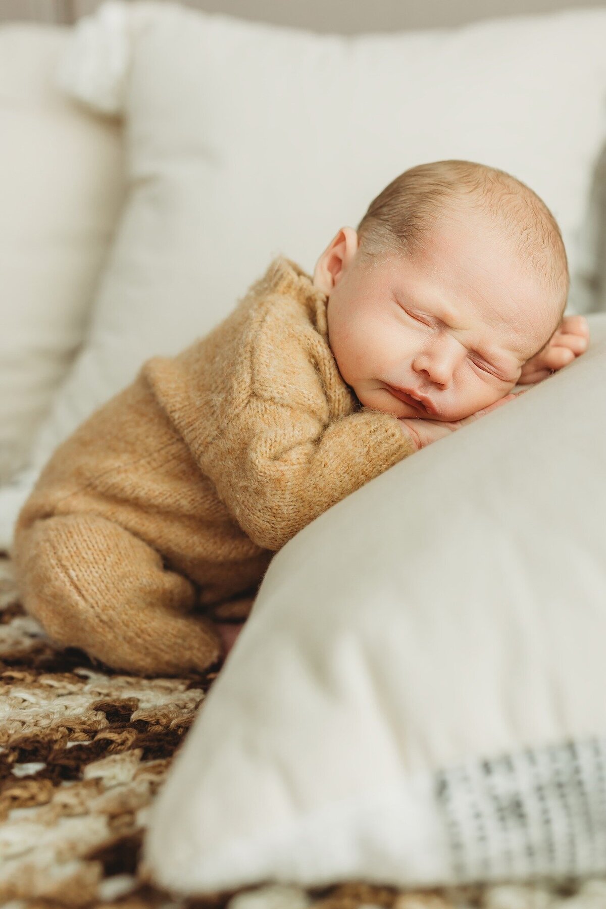 A newborn baby lies peacefully on a white pillow, wearing a soft tan outfit. The baby's eyes are closed, and they are curled up on their side, creating a serene atmosphere. The background features a cozy, textured blanket and additional pillows, enhancing the warmth of the scene.