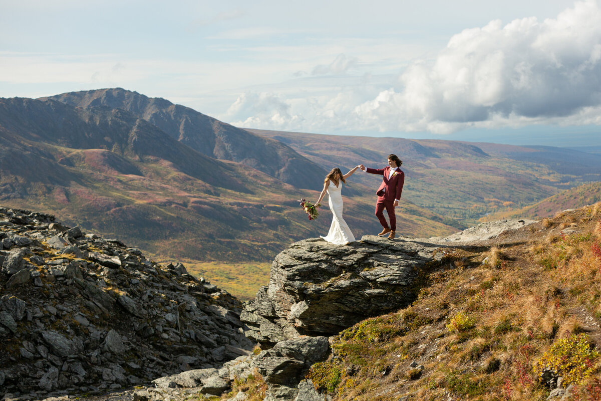 Bride in a sleek white dress and groom in a burgundy suit balance on a rock cliff, holding hands  