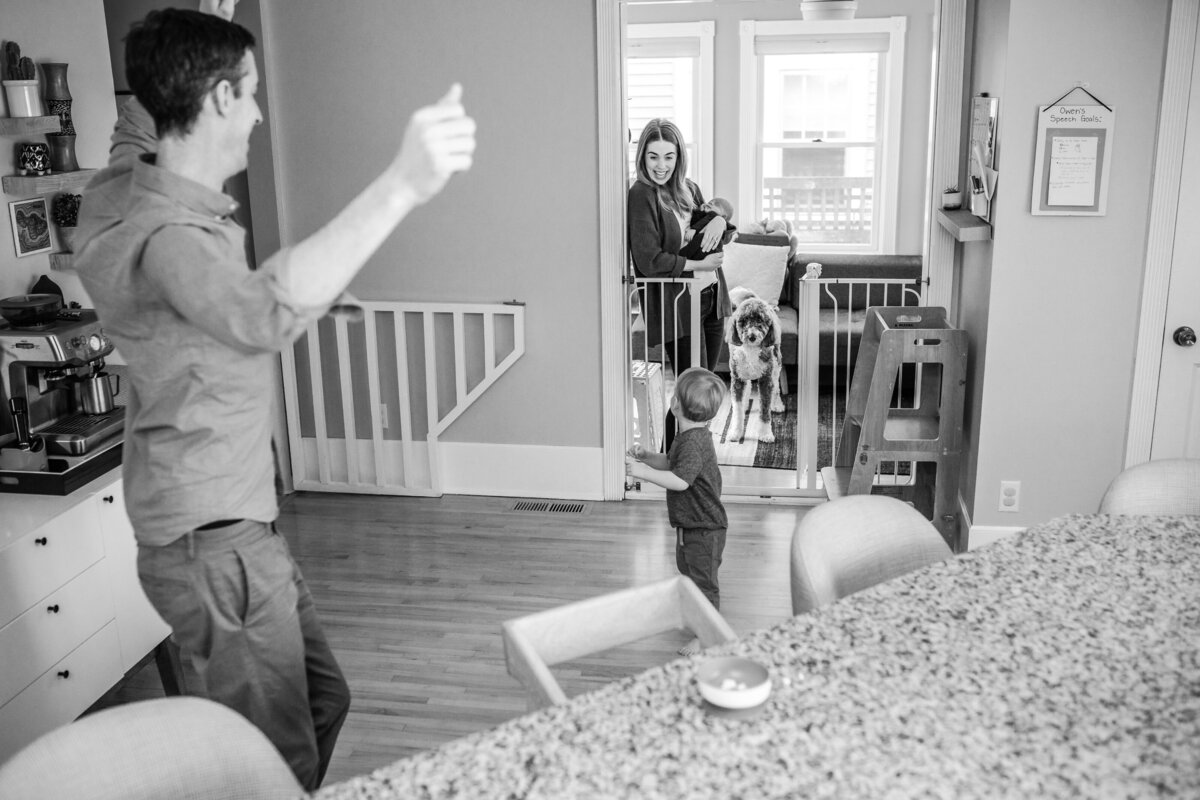 Black and white image of a father standing with his arms raised, greeting his toddler who is running towards him, while the mother stands in the background holding their newborn baby and watching, with a dog nearby.