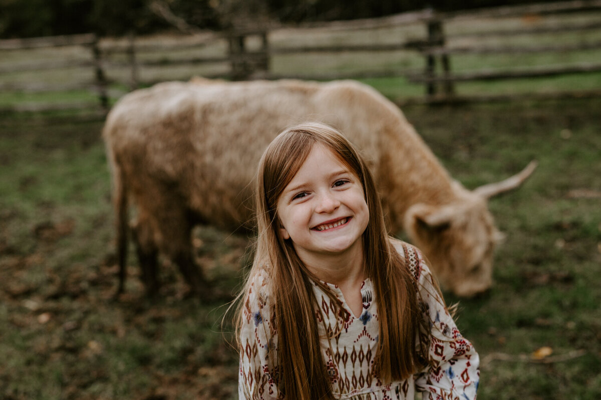Top family photographer shoot with highland cattle outside of London, Ontario. Portrait of a smiling girl with a baby highland cow behind her.