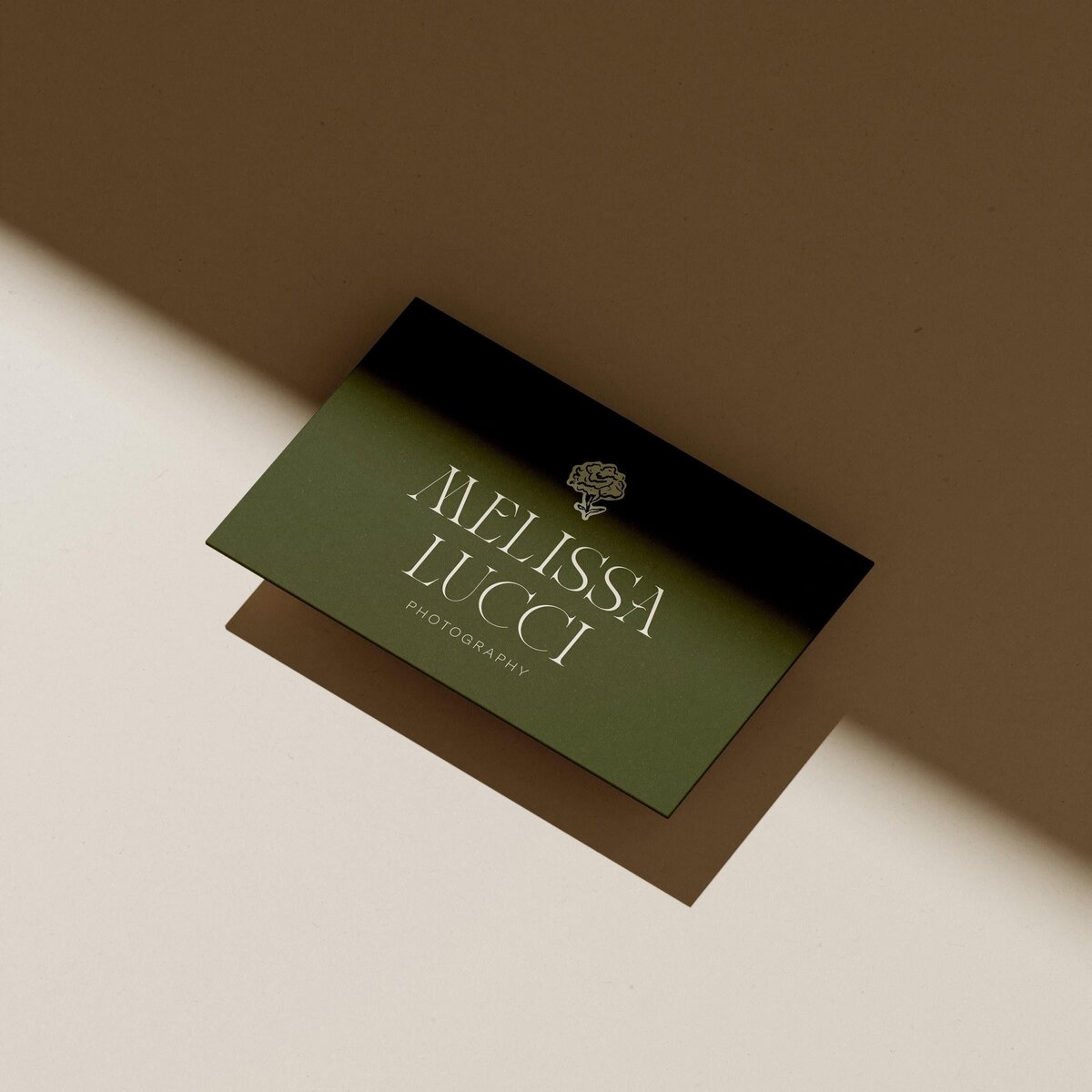 A business card with the text "MELISSA LUCCI PHOTOGRAPHY" and a small flower logo, designed by experts in brand strategy consulting, placed on a beige surface with a shadow cast over it.