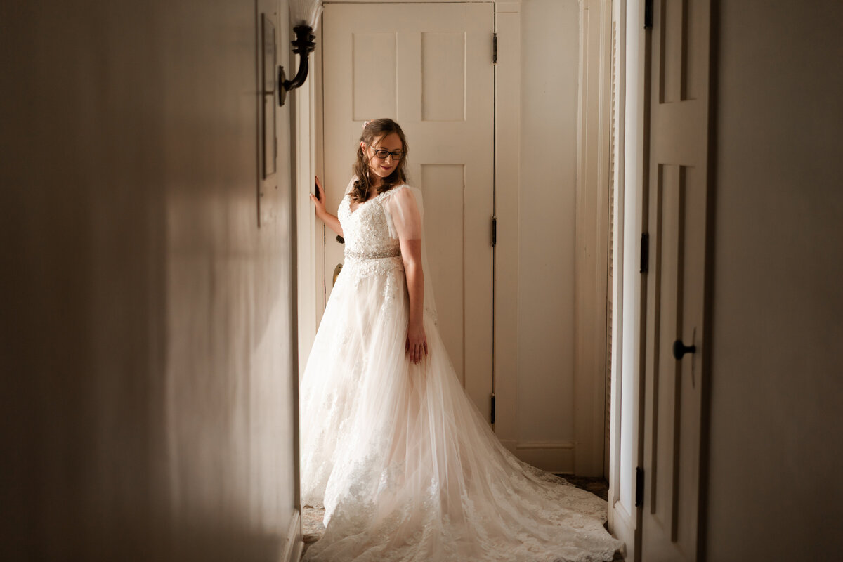 Bride stands in the window light and looks down off her shoulder in a hallway at her wedding at the Akron Woman's City Club.