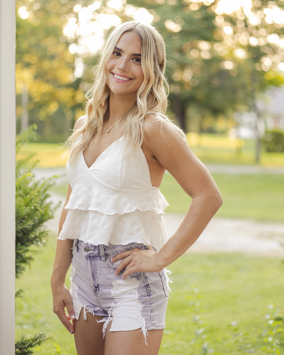 high school senior girl standing on a porch with long blonde hair,  a hand on her hip, slightly turned to the side smiling at the camera, wearing blue jean shorts, and spaghetti strapped white shirt 