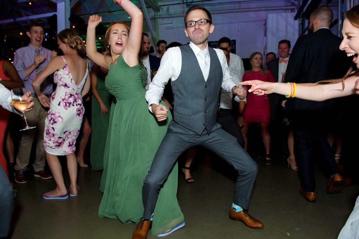 a groom dances silly with a bridesmaid as the bride looks on and laughs