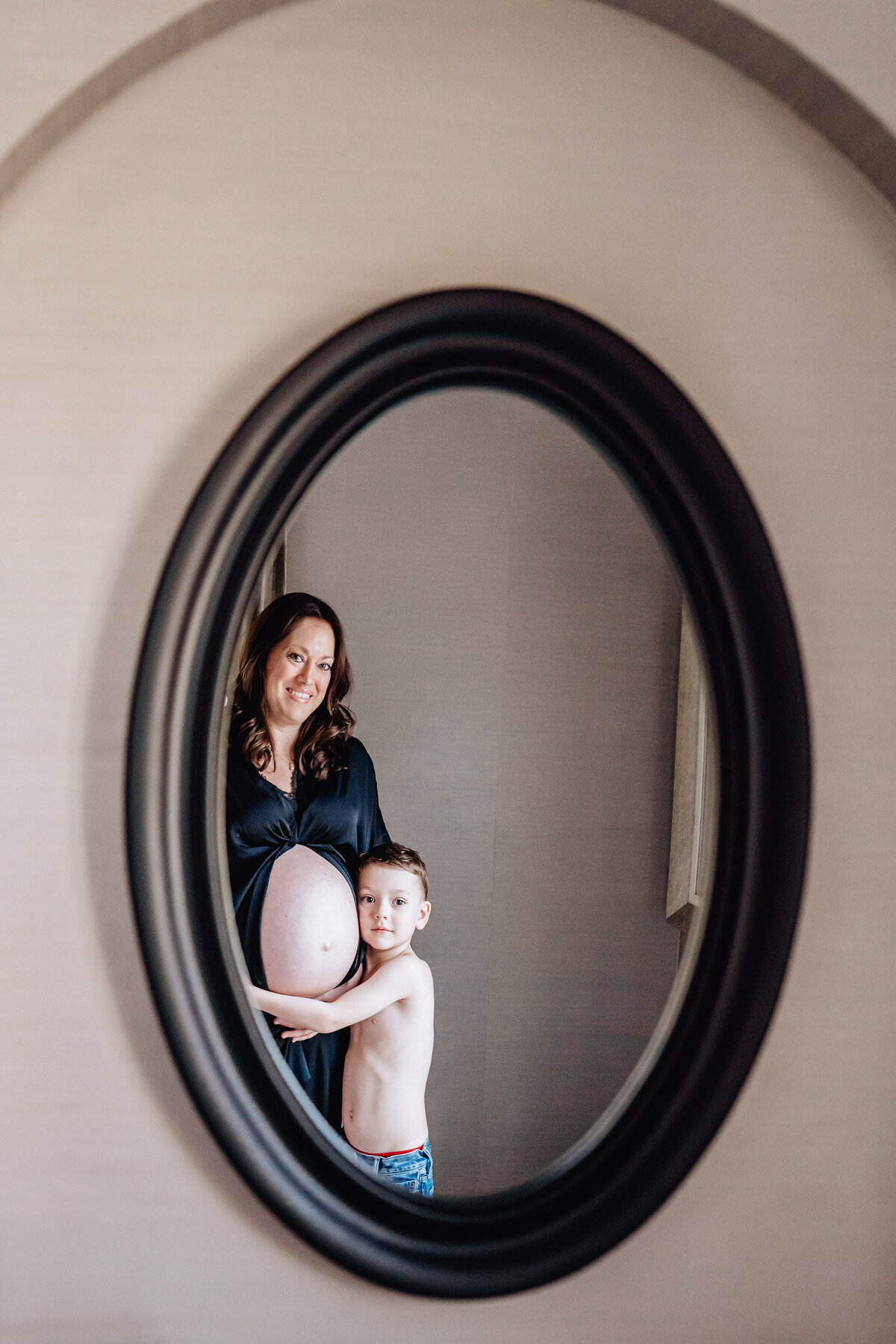 A pregnant woman and her young son are reflected in an oval mirror. She cradles her bare belly while her son, standing in front of her, hugs her with a gentle smile.