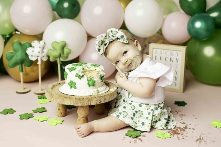 First birthday photoshoot with a baby girl dressed in a lucky-themed outfit, sitting on a bed of four-leaf clovers