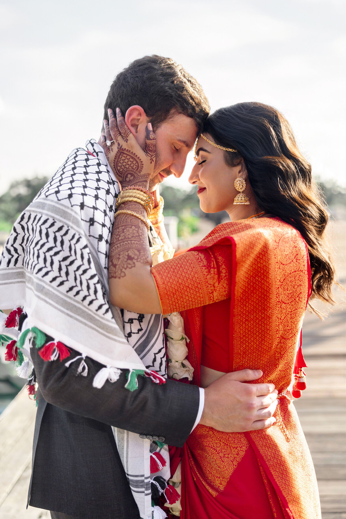 A couple in traditional attire embraces on a wooden path. The woman, in an orange sari with intricate henna designs on her hands, faces the man wrapped in a patterned scarf. Their foreheads touch gently, expressing affection.