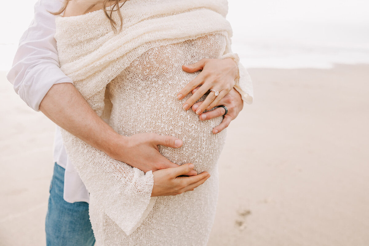 picture of a husband and wife's hands on pregnant belly. She is wearing an off the shoulder cream dress and the ocean is behind them