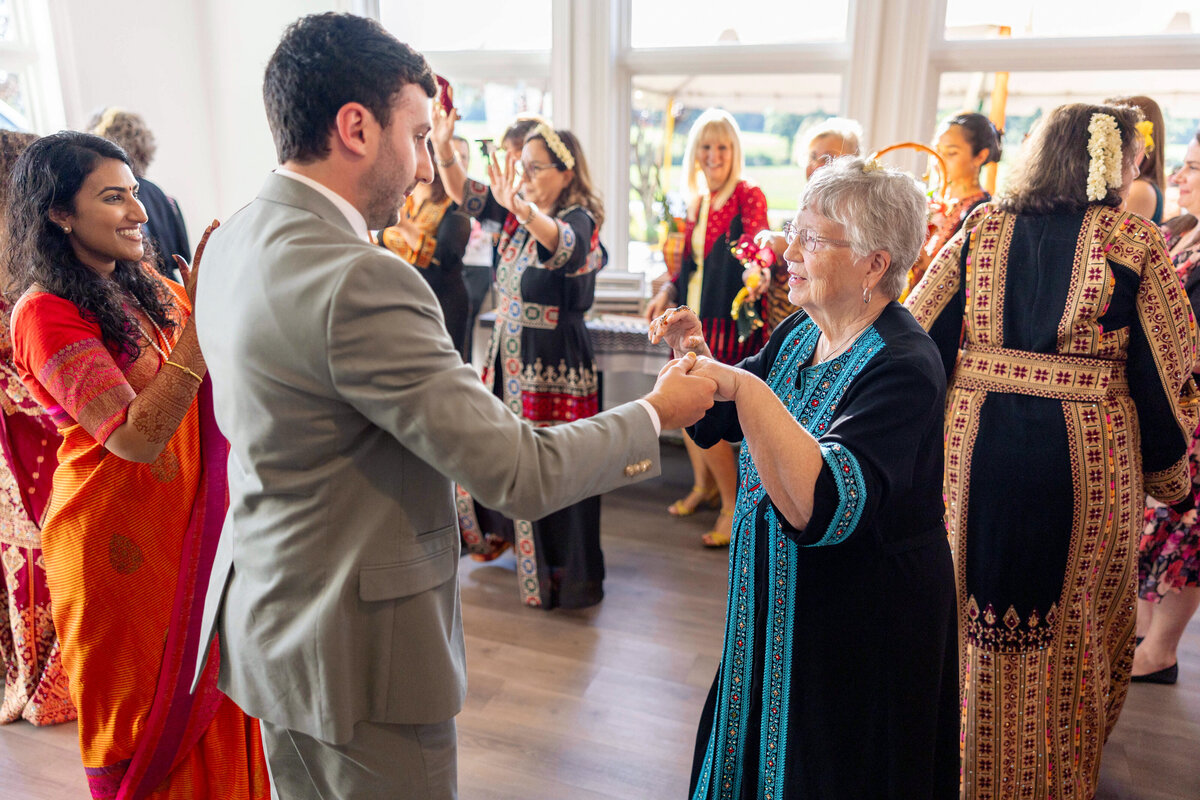 People dance joyfully at a festive gathering. A man in a suit and an older woman in a black and blue outfit hold hands, surrounded by others in colorful traditional attire, creating a lively and celebratory atmosphere.