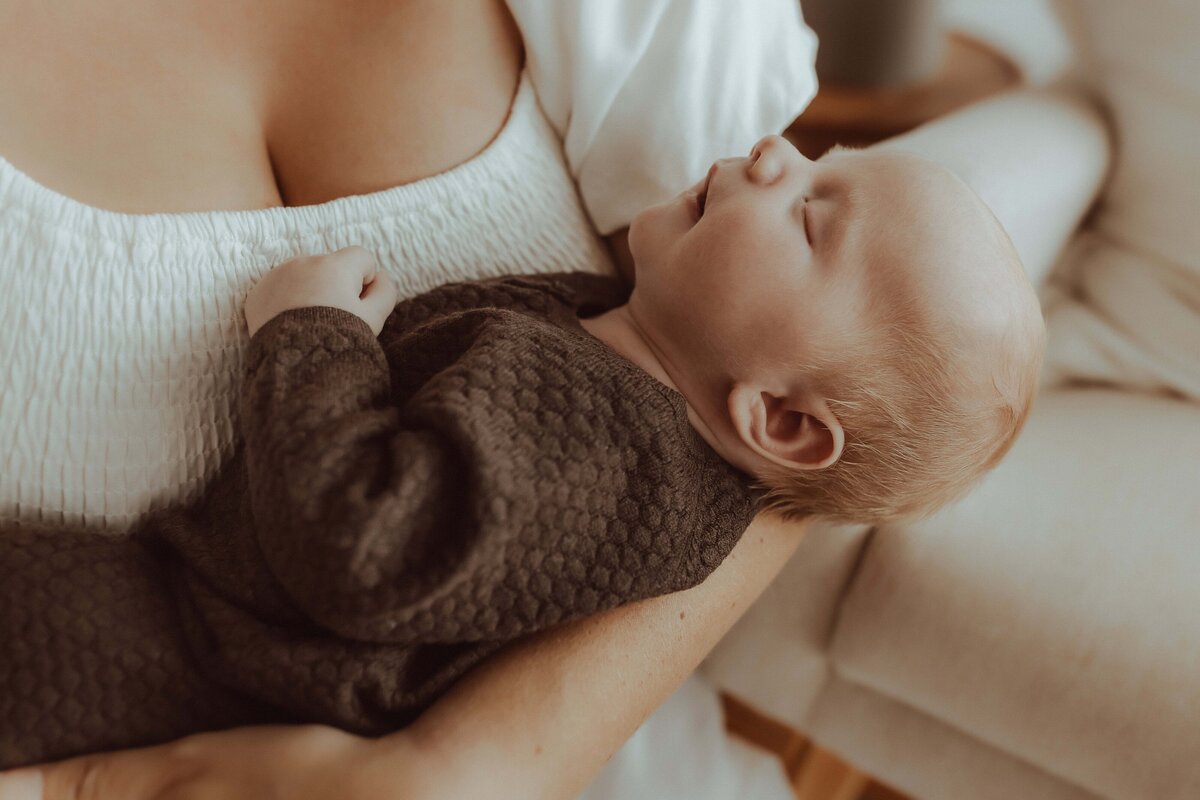A baby sleeps peacefully in a woman's arms. The baby is wearing a brown textured outfit. The woman is wearing a white top, and they are sitting on a cream-colored couch.