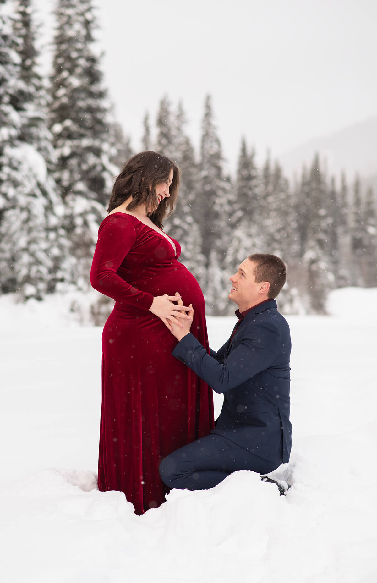 A couple is photographed at Sun Peaks for a maternity session. A woman wears a red velvet dress while looking at her husband who is crouched down holding her belly.