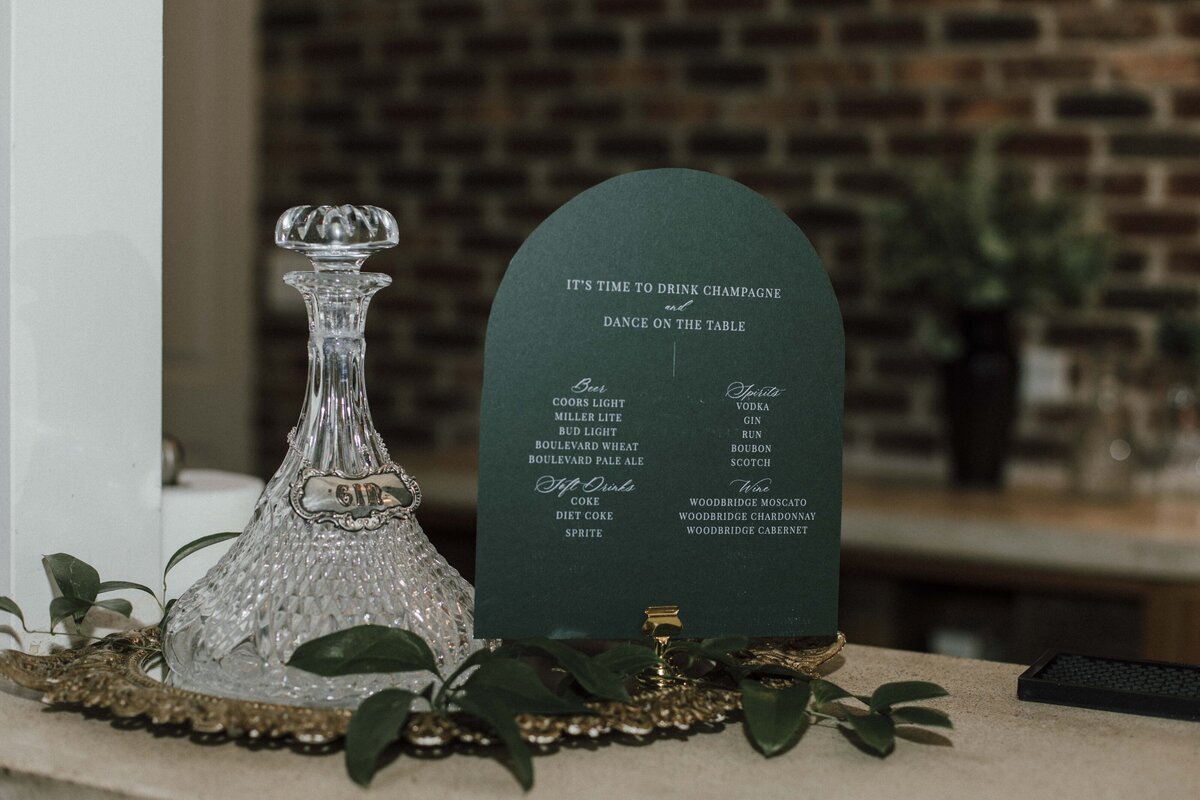 Rounded place card with white font on a gold place card holder atop a gold tray with leaves and a glass bottle.