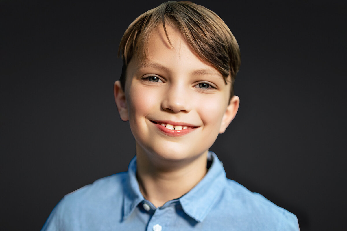 A 10 year old boy wearing a blue shirt with bright blue eyes smiles at the camera during a photo shoot at the Pittsburgh photography studio standing on a striking black background