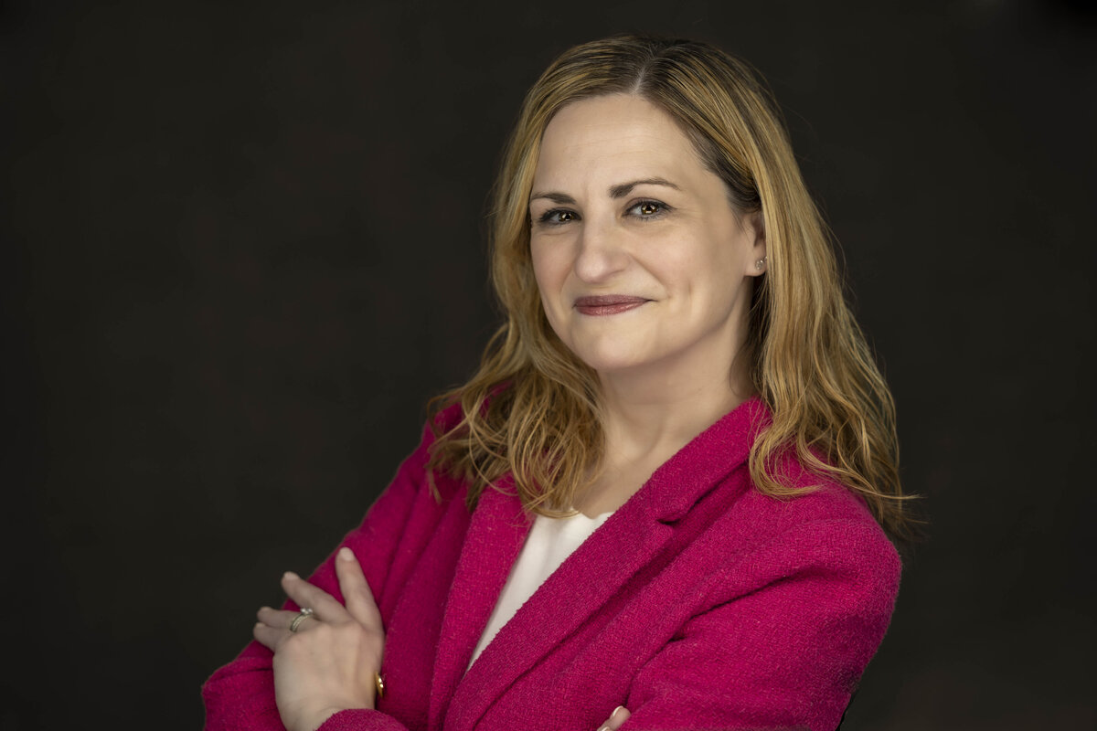 Professional headshot in CT of a woman in a vibrant pink blazer, arms crossed, smiling warmly in front of a dark textured background, by Rob Faber Photography, offering headshots in CT and headshots in MA.