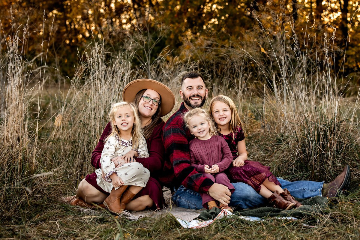 A family of five sits together on a blanket in a grassy field. The mother, wearing a hat, and father, in a plaid shirt, are surrounded by three young girls. The background features tall grass and trees with autumn foliage. Everyone is smiling.