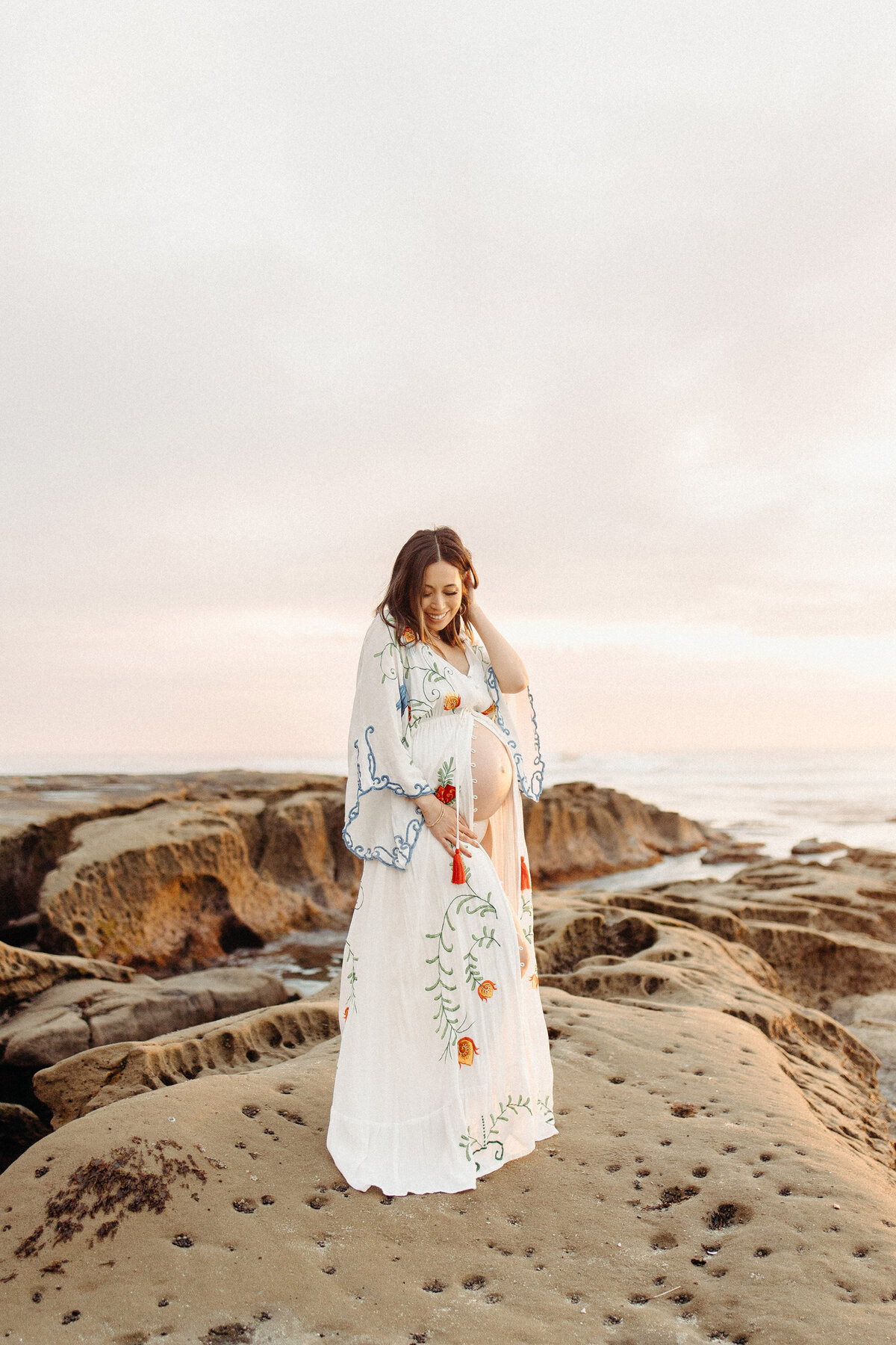 Beautiful maternity photoshoot at La Jolla beach with a mother-to-be in a white embroidered gown, standing on the rocky shore at sunset. Captured by San Diego Family Photographer Yuka Schad.