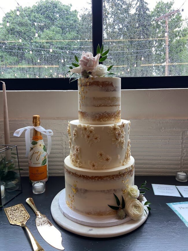 A three tier wedding cake sits in front of a window. The top and bottom of the cake have naked style frosting with gold leaf accents. The middle tier is frosted with white frosting and is decorated with a smattering of tiny buttercream flowers in white and very pale pink. On the top and at the bottom of the cake are two small clusters of fresh flowers and greenery.