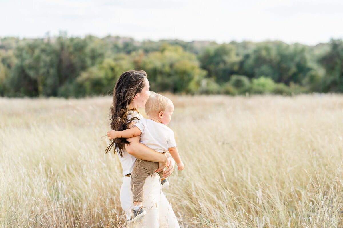 a mother stands with her toddler son in her arms in a field of golden, tall grass and looks away from the camera of her Austin family photographer.
